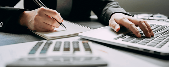 Close-up of hands writing in a notebook beside a calculator and laptop