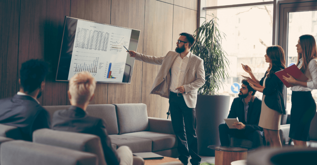 Business team gathered in a modern office presenting financial data on a wall-mounted screen showing charts and graphs, with team members reviewing documents while listening to a male presenter.